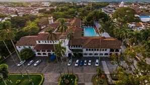 An aerial view of a residential area with a large building featuring a swimming pool, surrounded by palm trees and parked cars.