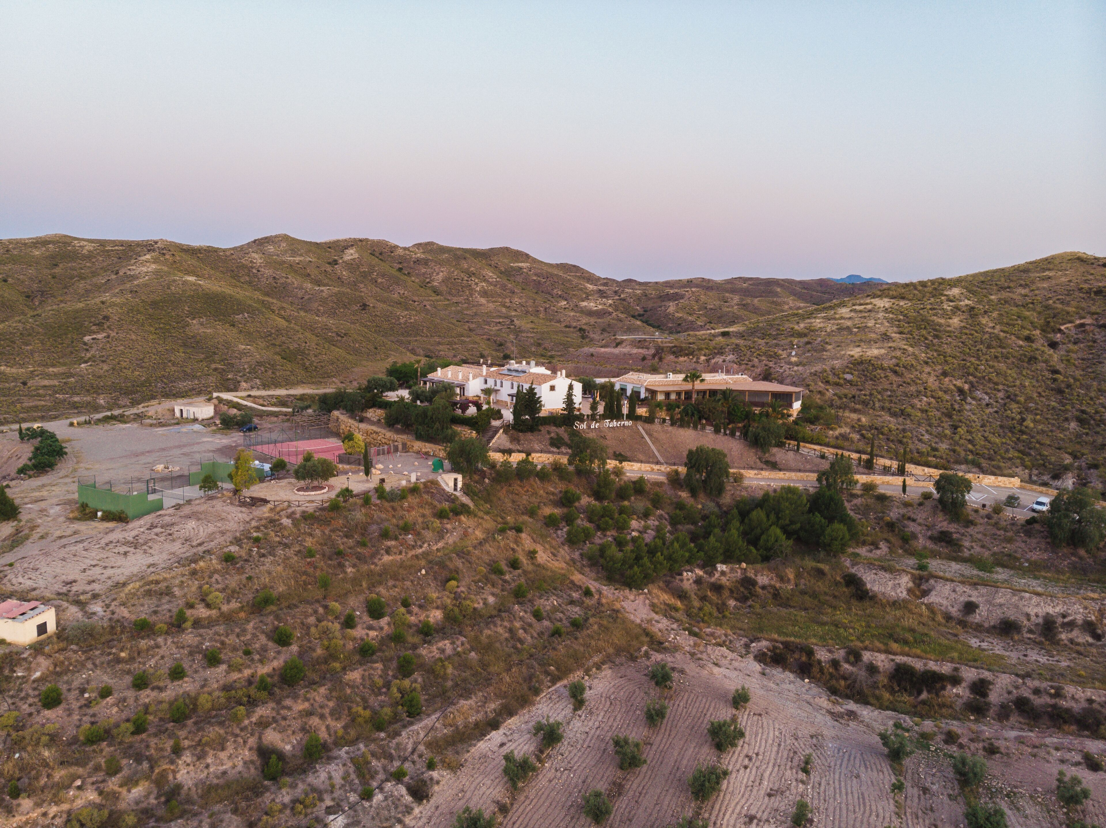 A large, white building with multiple sections, surrounded by a hilly landscape.