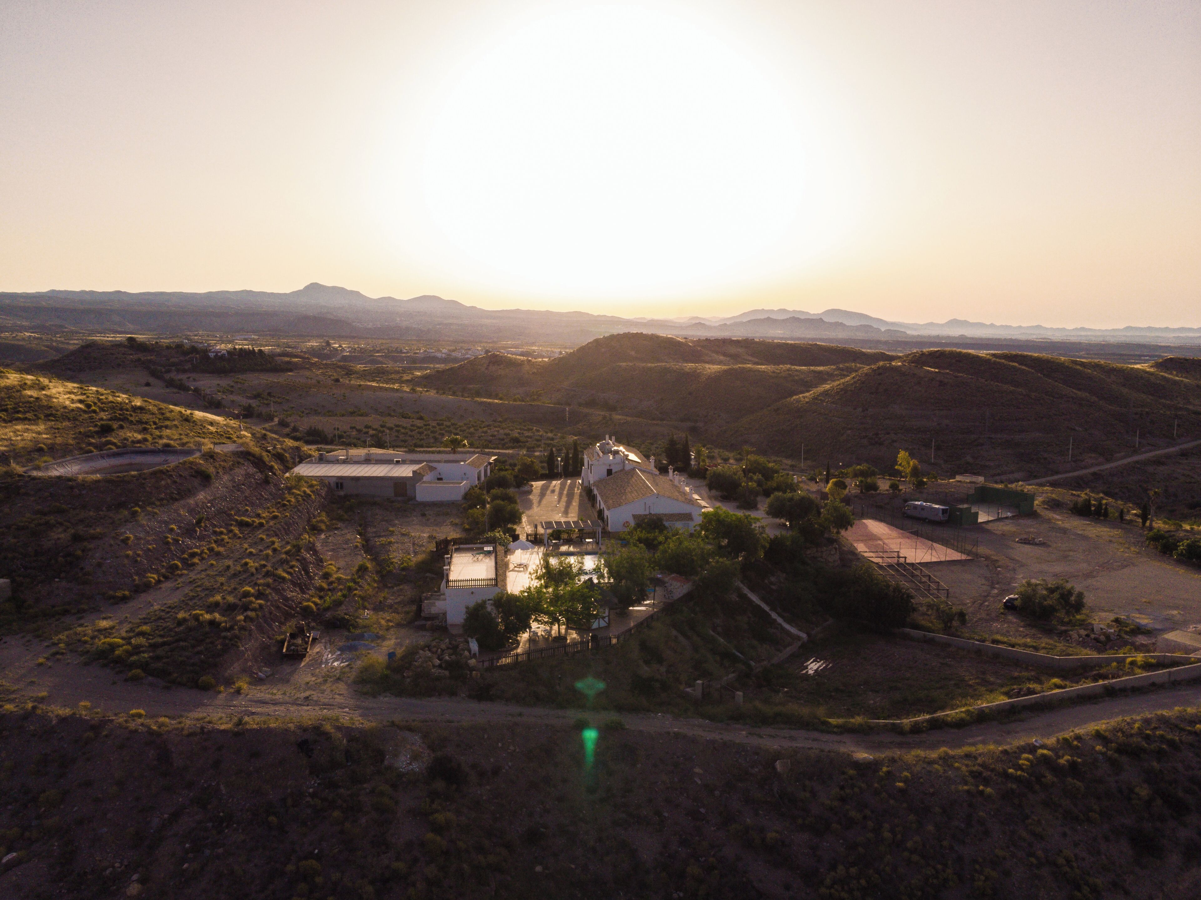 Un paisaje rural con un pequeño grupo de construcciones, incluyendo una casa blanca de techo rojo, rodeado de colinas y vegetación escasa.