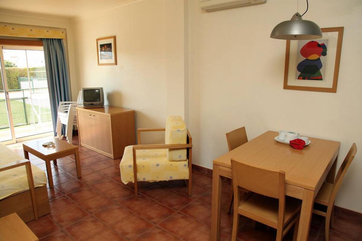 A room with a dining table, chairs, a small cabinet, a television, and a window with curtains.