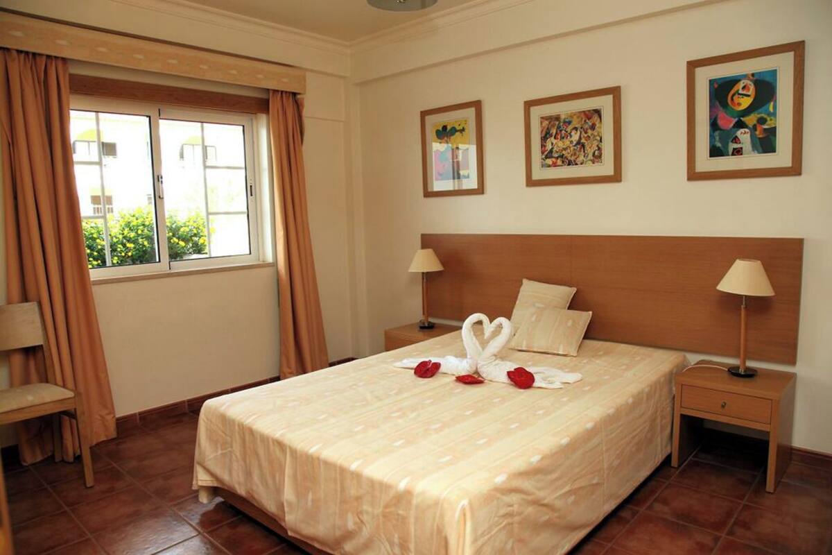 A hotel room with a large bed, two bedside tables, a window with curtains, and three framed artworks on the wall.
