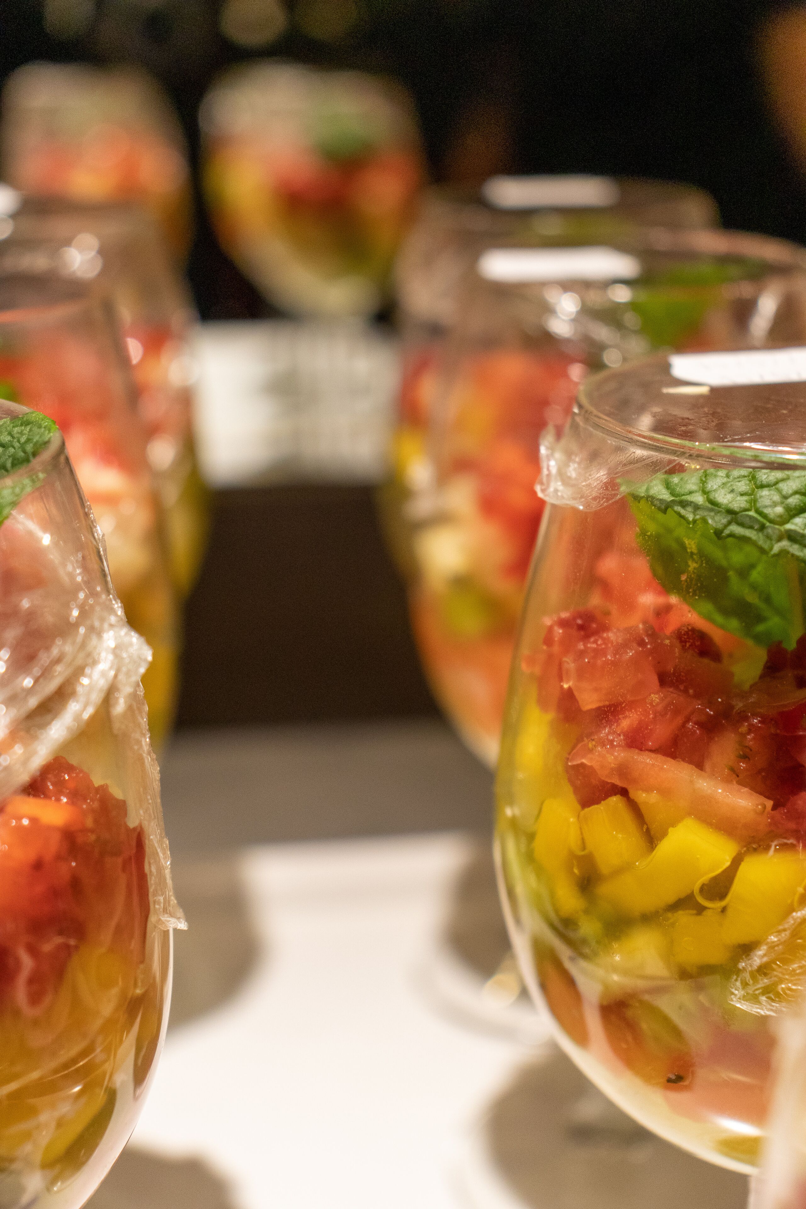 A close-up of a glass containing a layered fruit and vegetable salad with mint leaves on top.