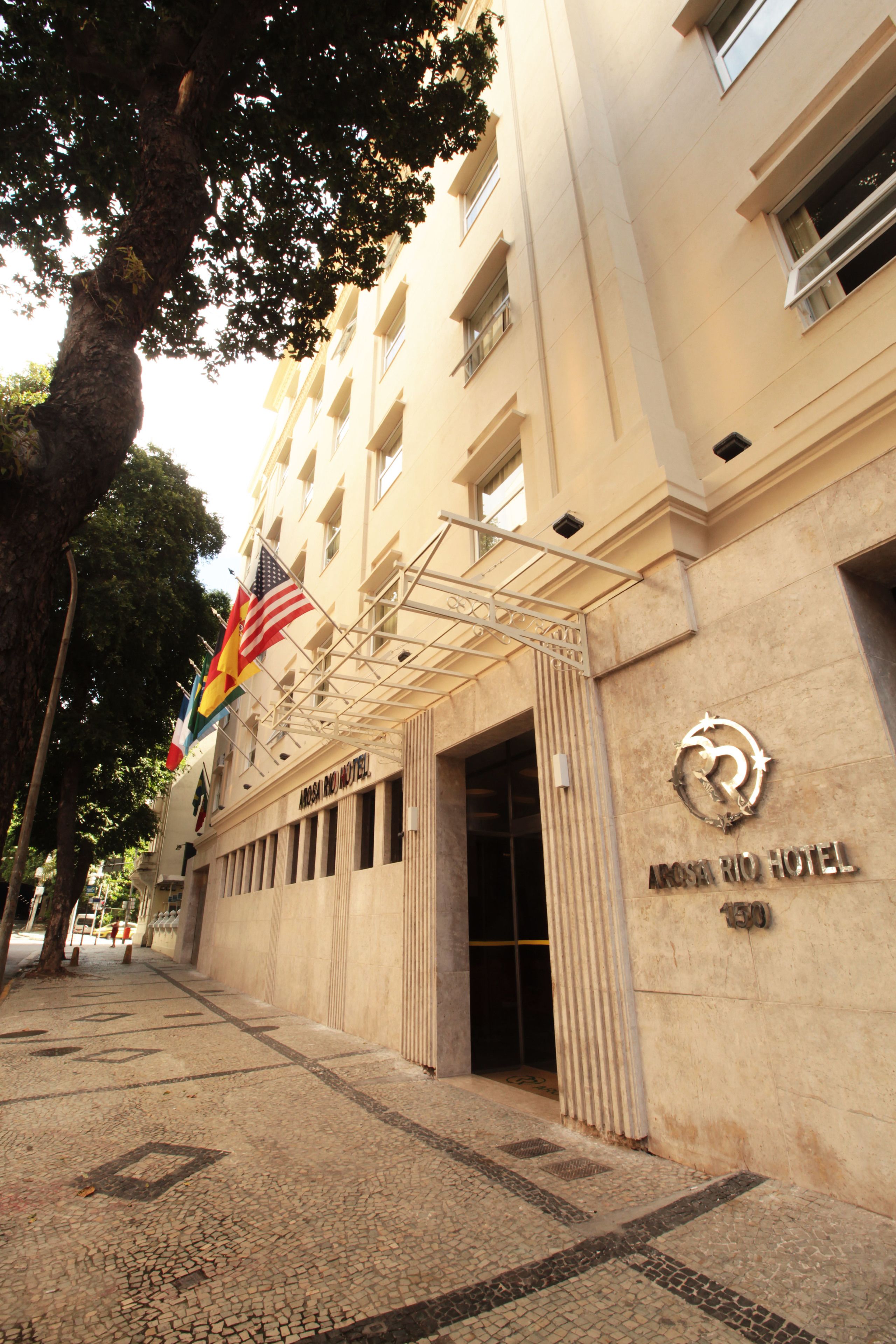 Exterior del Hotel Arcos Rio, con una entrada despejada, exhibición de banderas y un letrero de hotel llamativo.