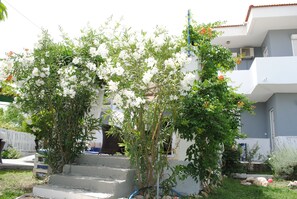 A residential building with a white exterior, a visible staircase leading to the entrance, and a garden area with white flowering plants and a small tree. - Karmik Concept Apartments (Rhodes)