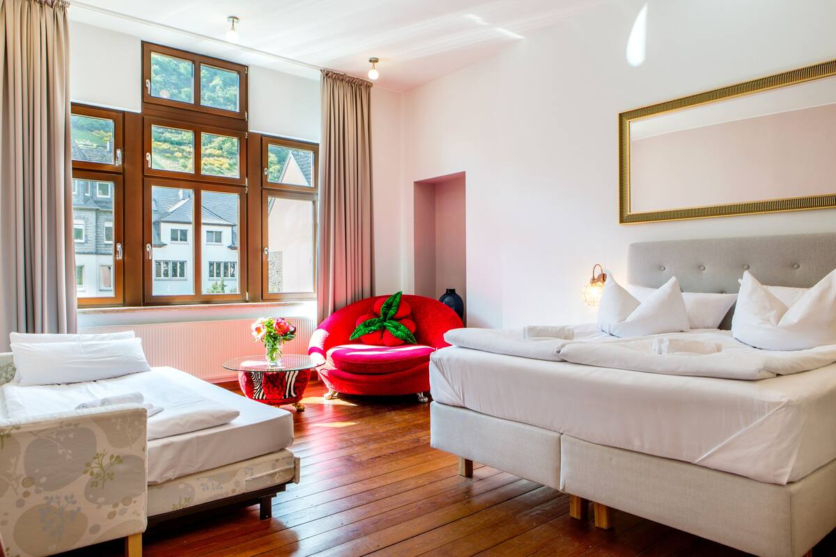 A hotel room with two beds, a red chair, a small table with a vase, and a large window with curtains.