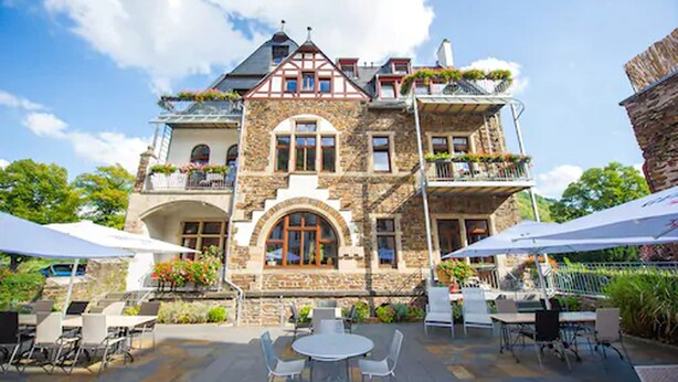 A stone building with a large arched entrance, multiple balconies with plants, and outdoor seating with umbrellas. - Hotel Villa Vinum Cochem (Cochem)
