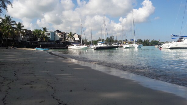 Ein Strand mit vor Anker liegenden Booten, sandigem Ufer und Gebäuden im Hintergrund.