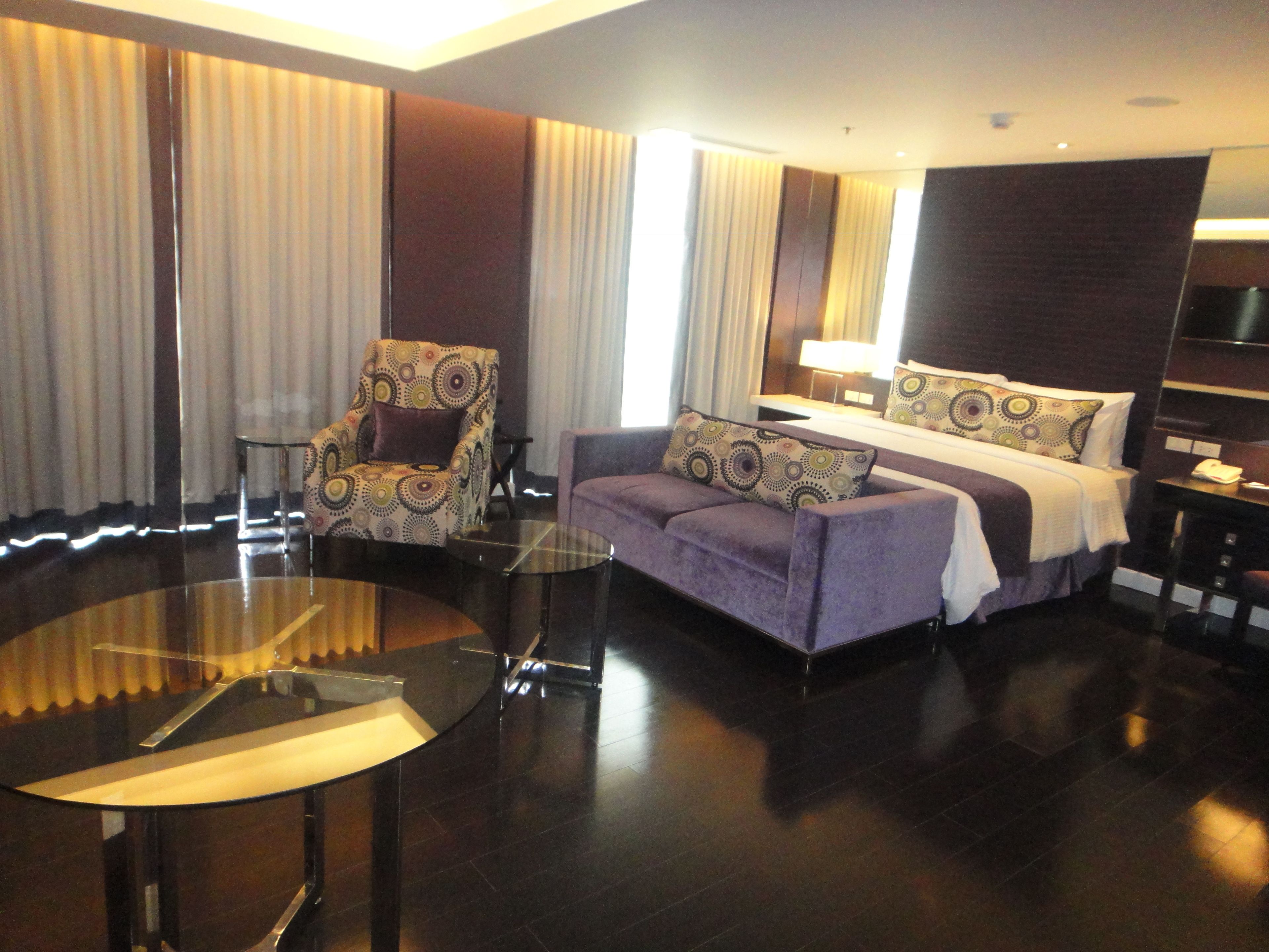 A modern hotel room with a bed, a sofa, a chair, a glass table, and a TV.