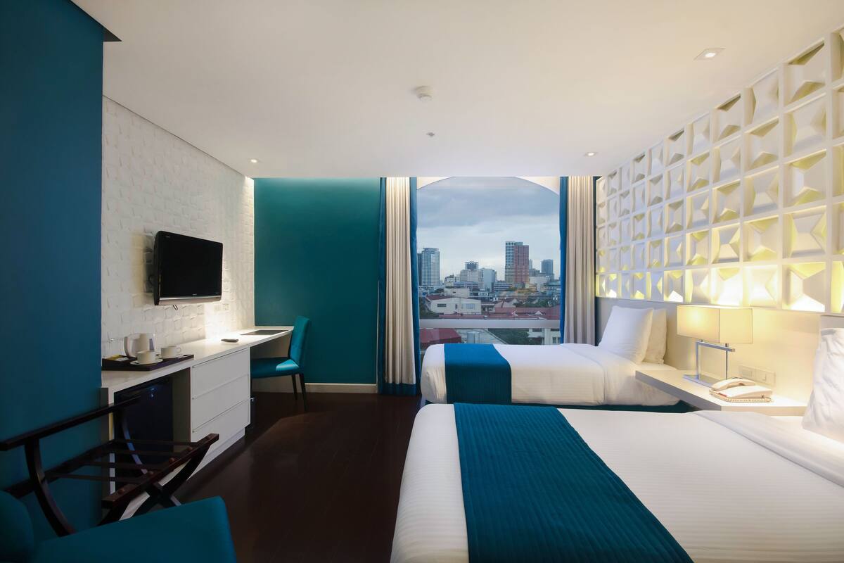 Ein Hotelzimmer mit Blick auf die Stadt, ein Schreibtisch mit Flachbildfernseher, ein Bett mit blauen Akzenten und einer wandmontierten Leuchte.