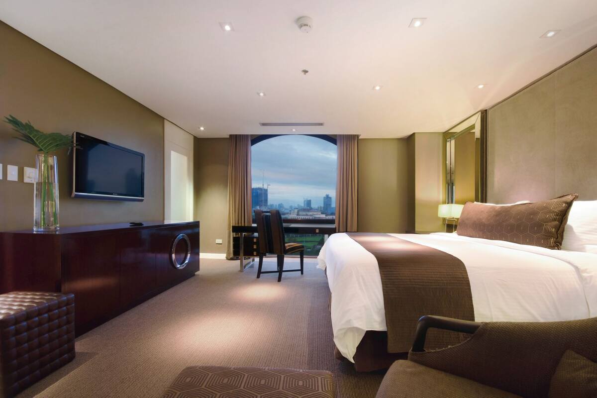 Ein Hotelzimmer mit einem großen Bett, einem Schreibtisch, einem Stuhl, einem Fernseher und Blick auf eine Stadtlandschaft.
