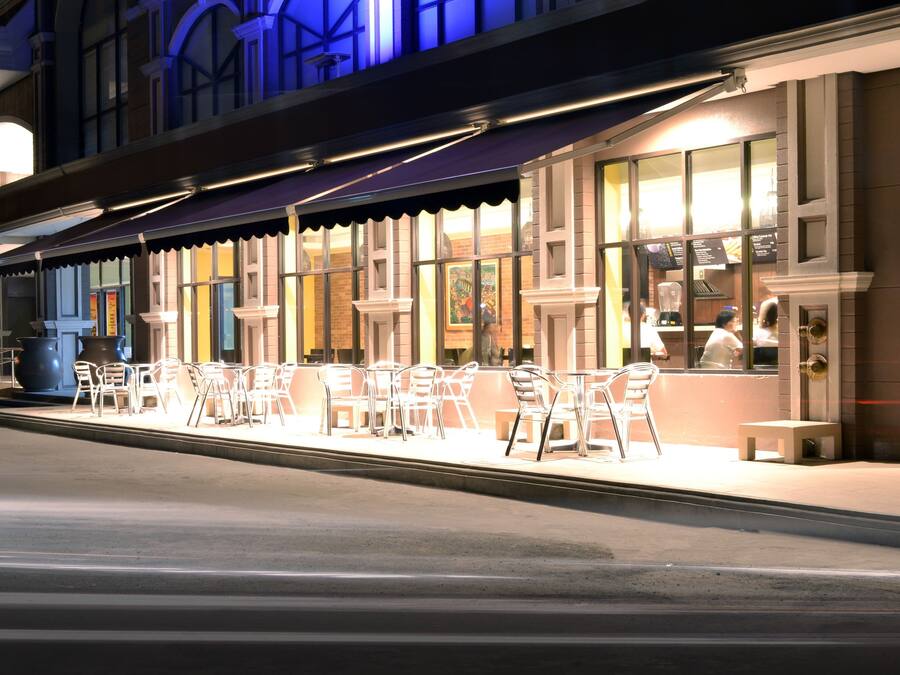 An outdoor seating area with tables and chairs, a building with large windows, and a street in front.
