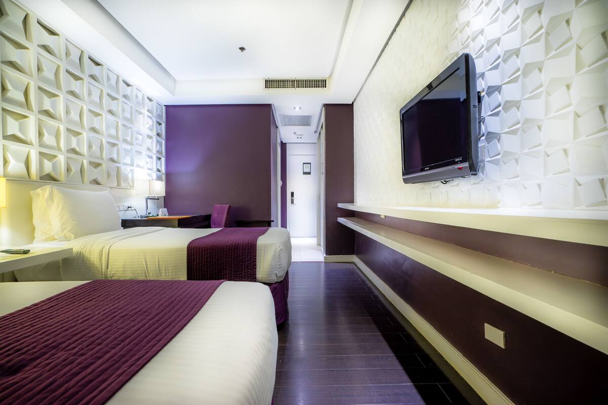 Ein Hotelzimmer mit zwei Betten, einem an der Wand befestigten Fernseher und einem Flur, der zu einem anderen Zimmer führt.