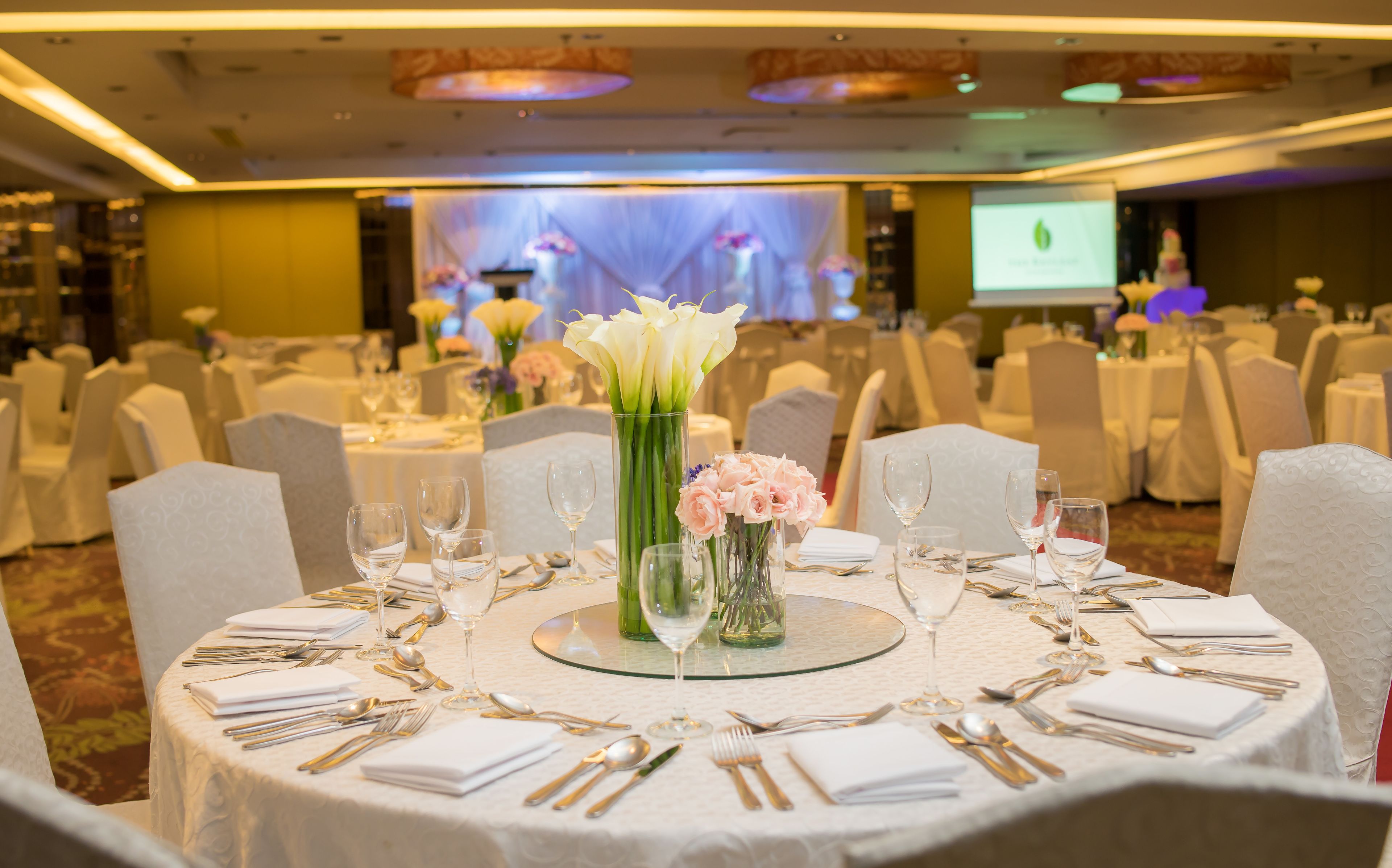 Un salón de banquetes con mesas redondas dispuestas para un evento formal, con manteles blancos, cubertería y centros de mesa florales.