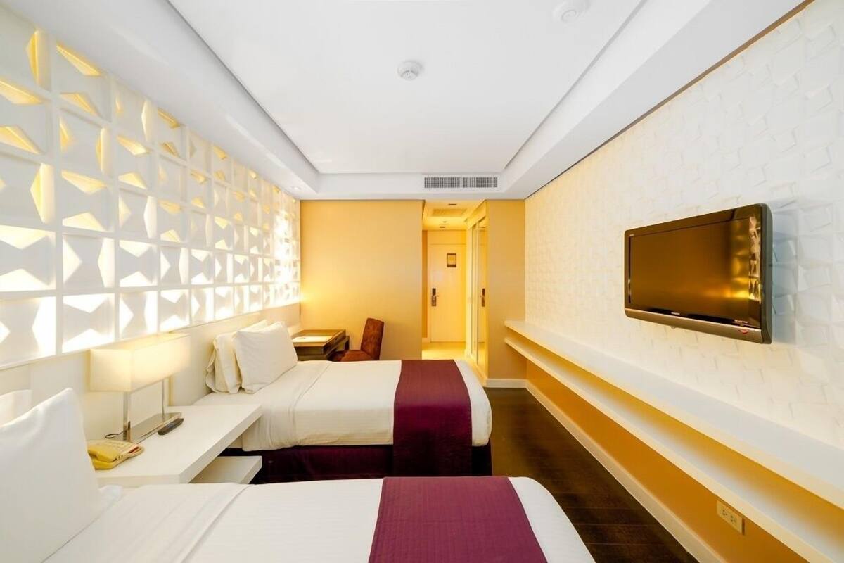 Ein Hotelzimmer mit zwei Betten, einem an der Wand befestigten Fernseher, einem Schreibtisch mit Lampe und einer Wand mit Muster.