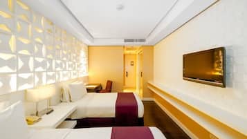A hotel room with two beds, a TV mounted on the wall, a desk with a lamp, and a patterned wall.
