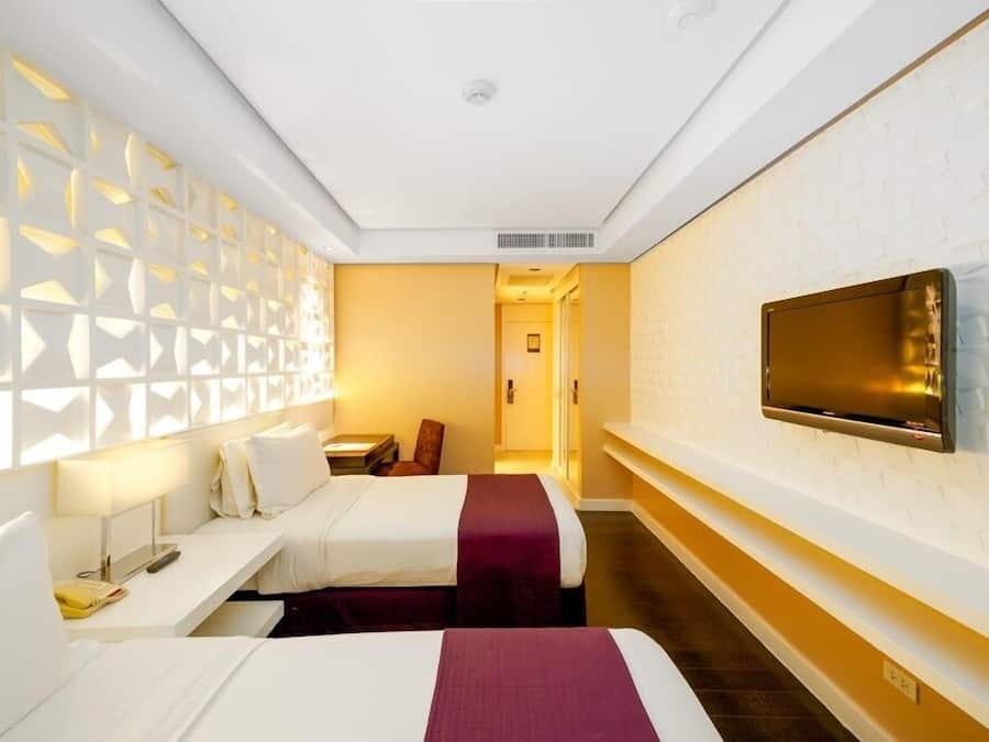 A hotel room with two beds, a TV mounted on the wall, a desk with a lamp, and a patterned wall.