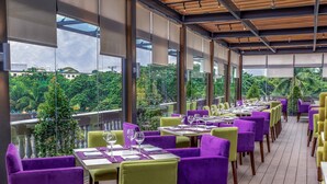 Un restaurante con sillas moradas y verdes, mesas blancas y amplios ventanales que ofrecen vistas a zonas verdes y edificios.