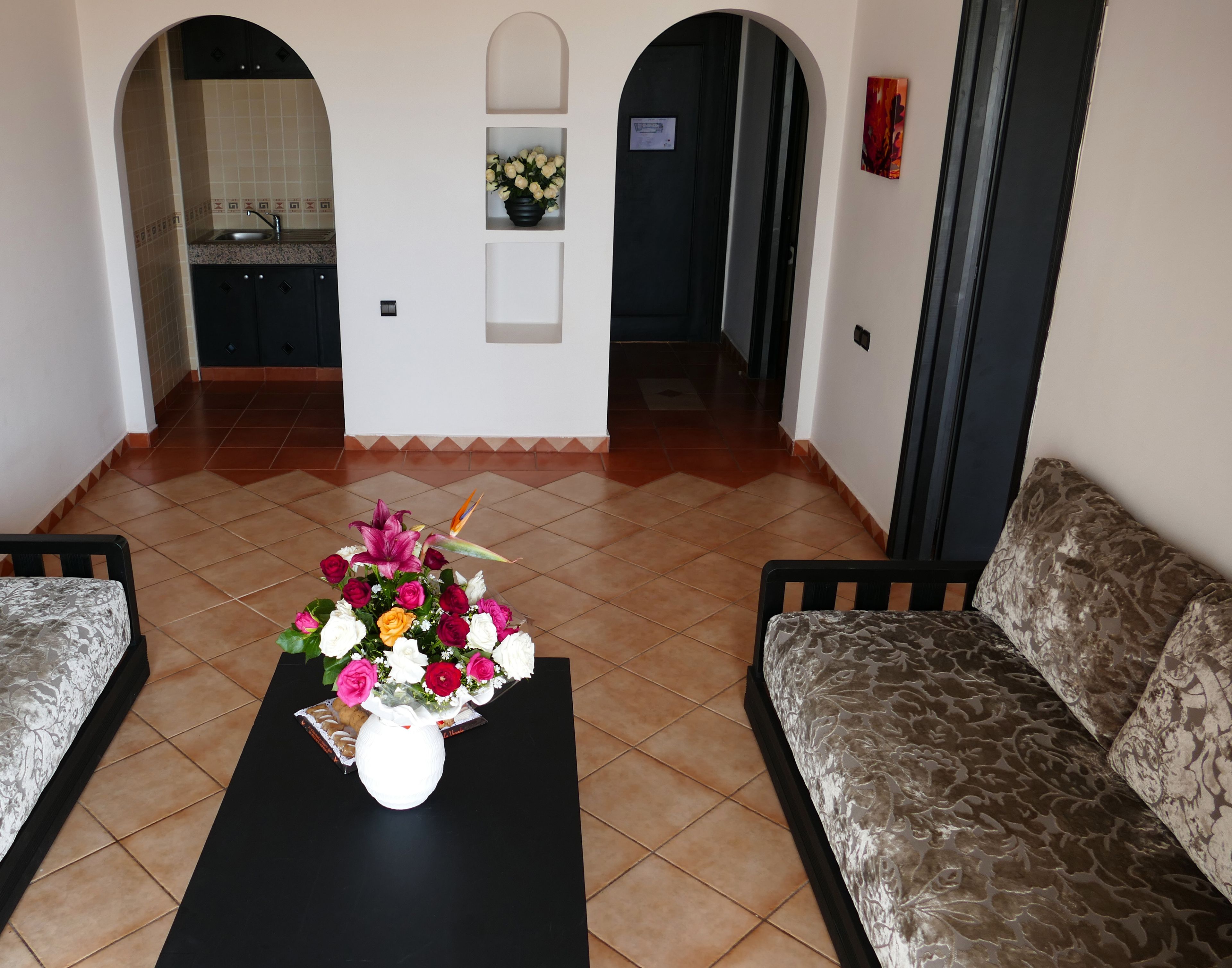 A living room with a black coffee table, floral arrangement, patterned sofa, and a kitchen area in the background.