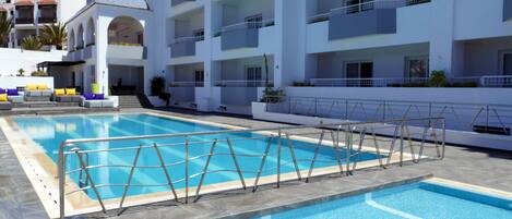 A swimming pool with a metal safety fence, surrounded by a white building with balconies and a covered seating area.