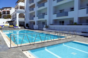 A swimming pool with a metal safety fence, surrounded by a white building with balconies and a covered seating area.