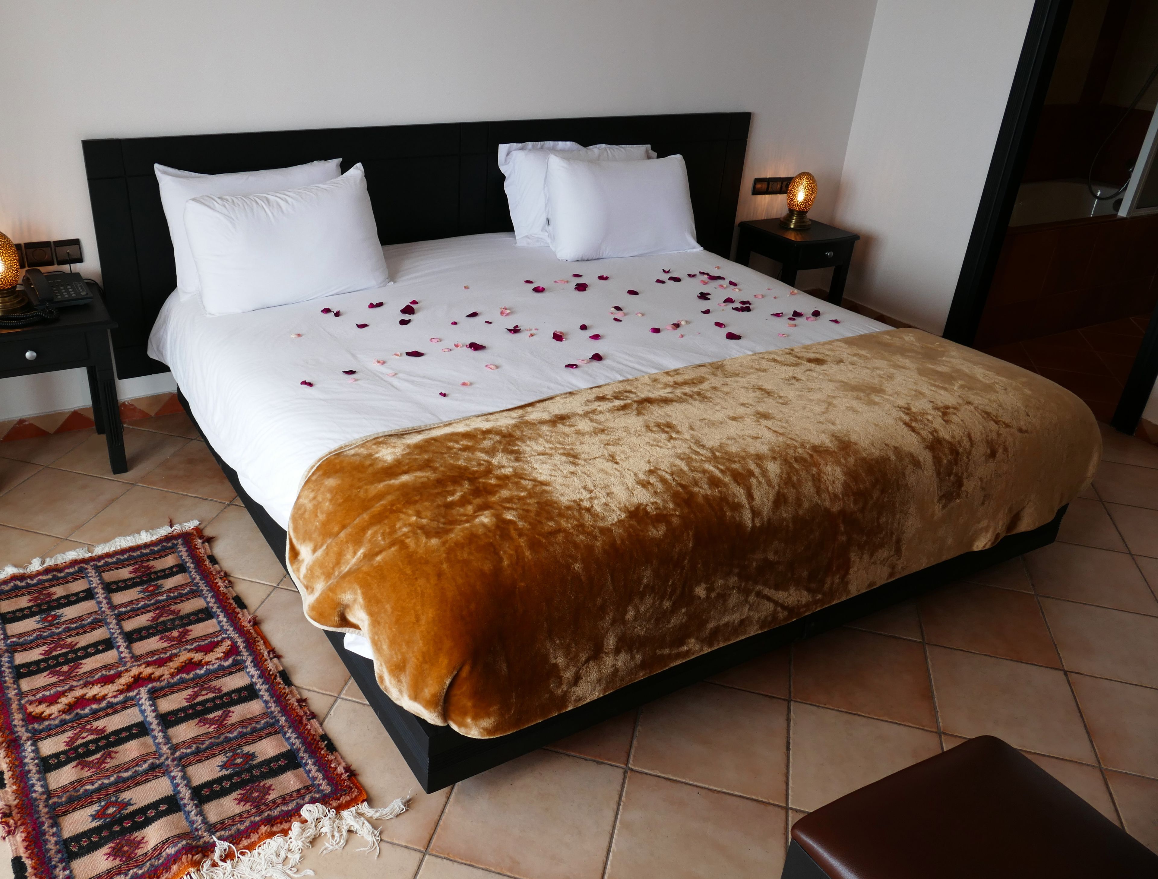 Una cama bien hecha con sábanas blancas y una manta marrón, un tapete estampado y una mesita de noche con una lámpara.