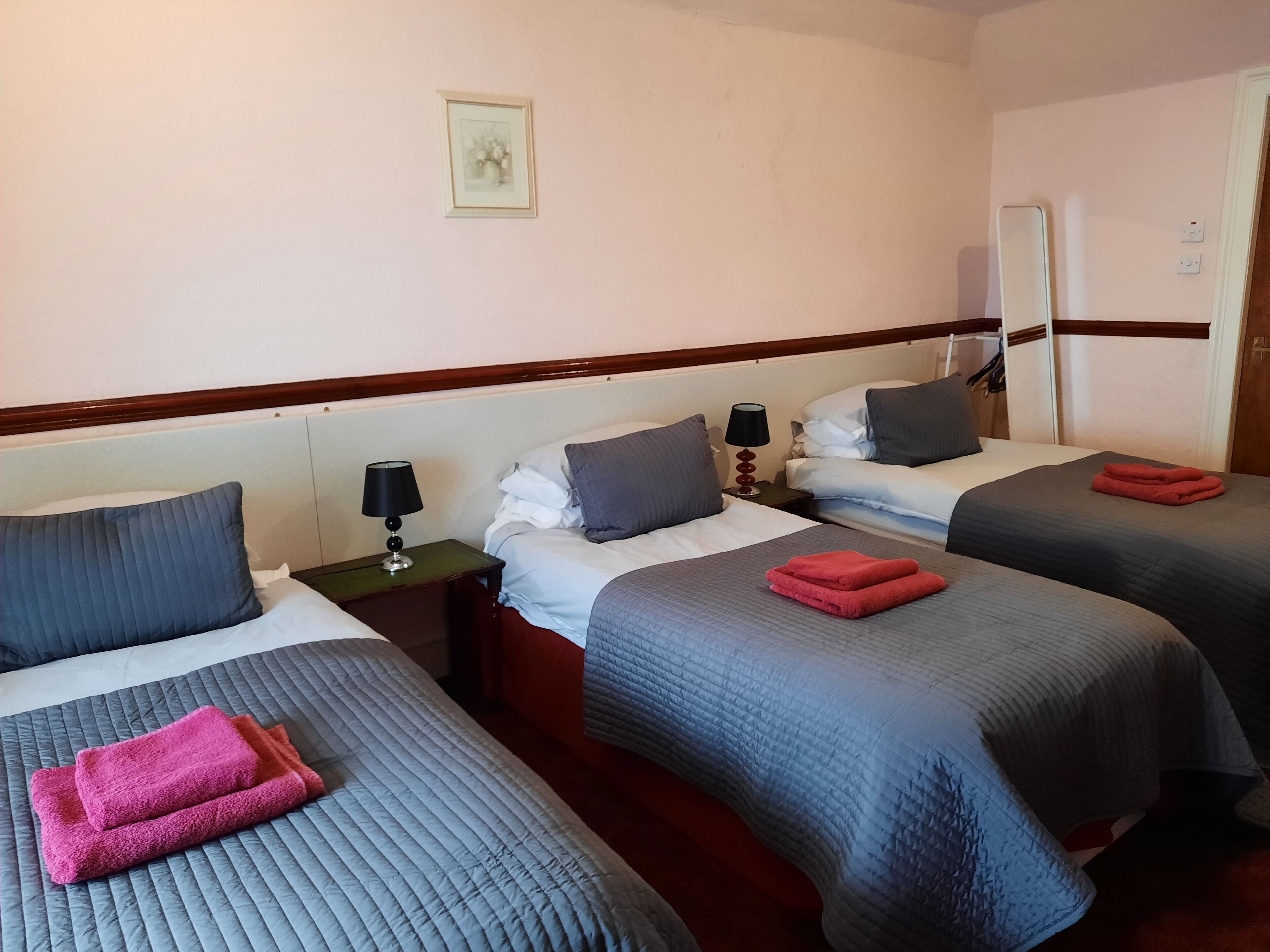 Habitación de hotel con tres camas, cada una con una manta y almohadas de colores diferentes. Hay una mesita con una lámpara y un cuadro en la pared.