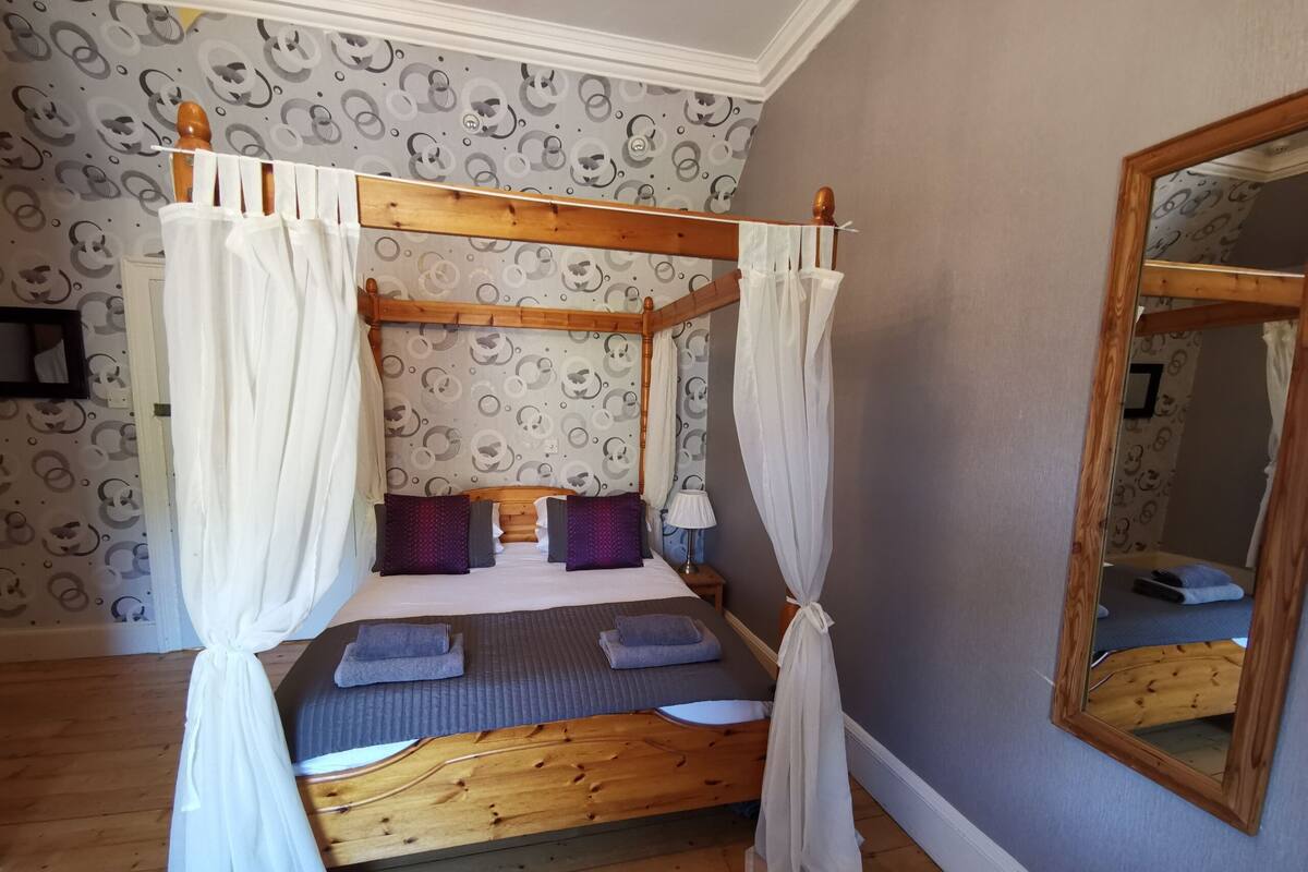 Una cama con dosel y cortinas blancas, cabecera de madera y papel tapiz estampado.