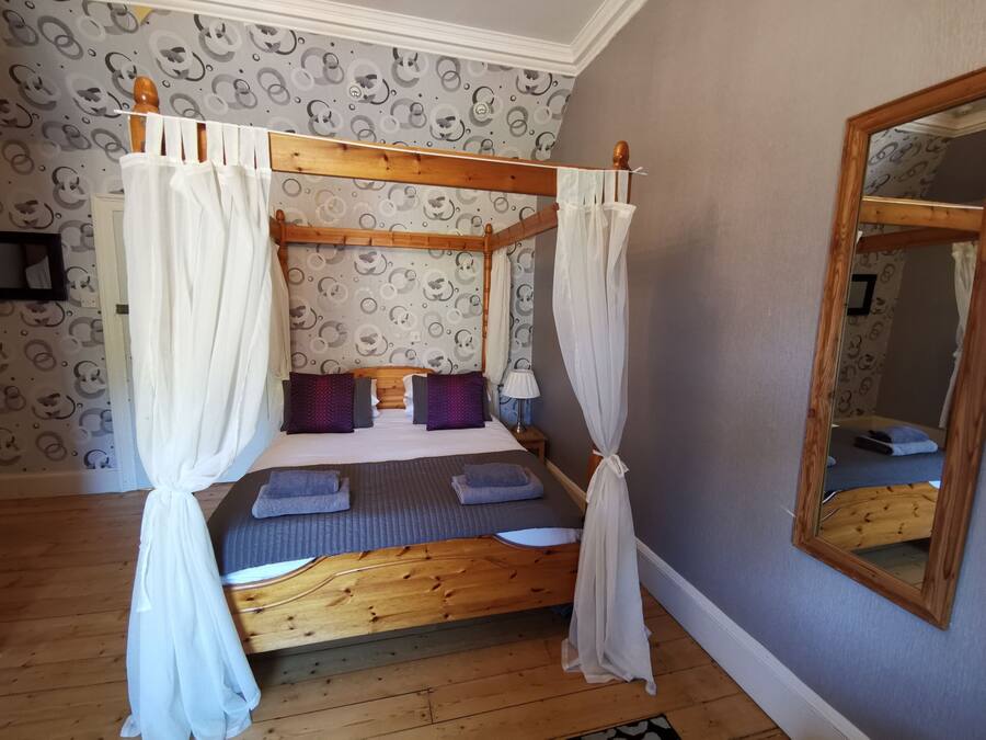 Una cama con dosel y cortinas blancas, cabecera de madera y papel tapiz estampado.