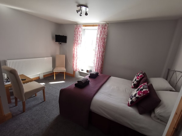 A hotel room with a bed, a desk with a chair, a TV, and a window with curtains. - St Ronans Hotel (Peebles)