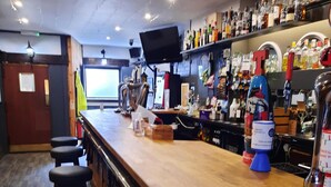 Una zona de bar con mostrador de madera, taburetes y una variedad de botellas y vasos en estantes.