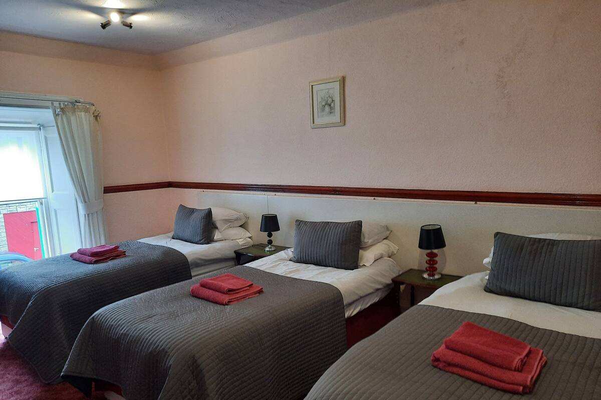 Habitación de hotel con tres camas individuales, cada una con una almohada y una toalla roja. Hay dos mesitas de noche con lámparas.