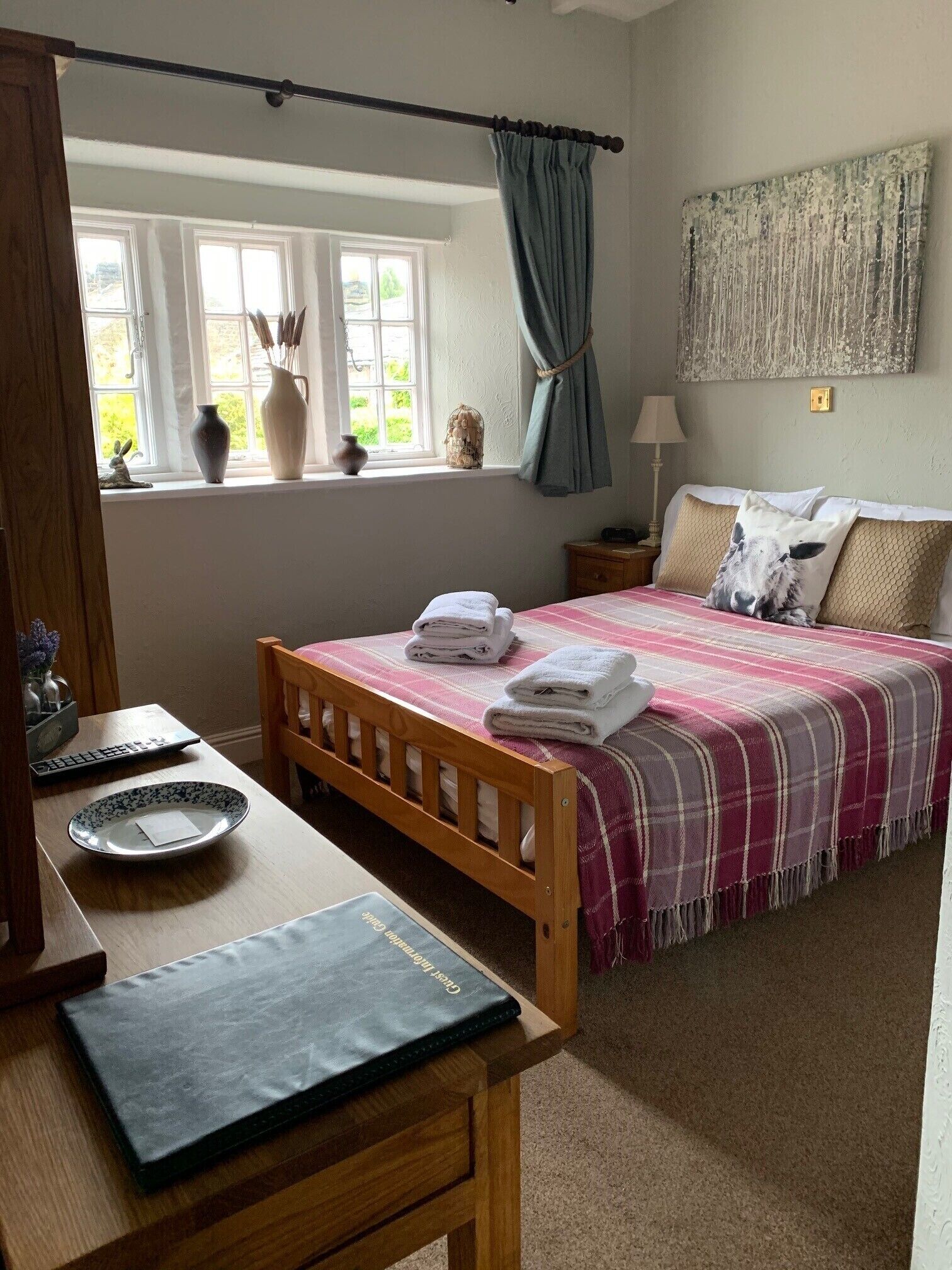 A bedroom with a wooden bed, a bedside table with a lamp, a window with curtains, and a view of greenery outside.
