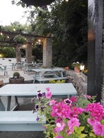 Un patio al aire libre con pilares de piedra, mesas de picnic de madera y cestas de flores colgantes.