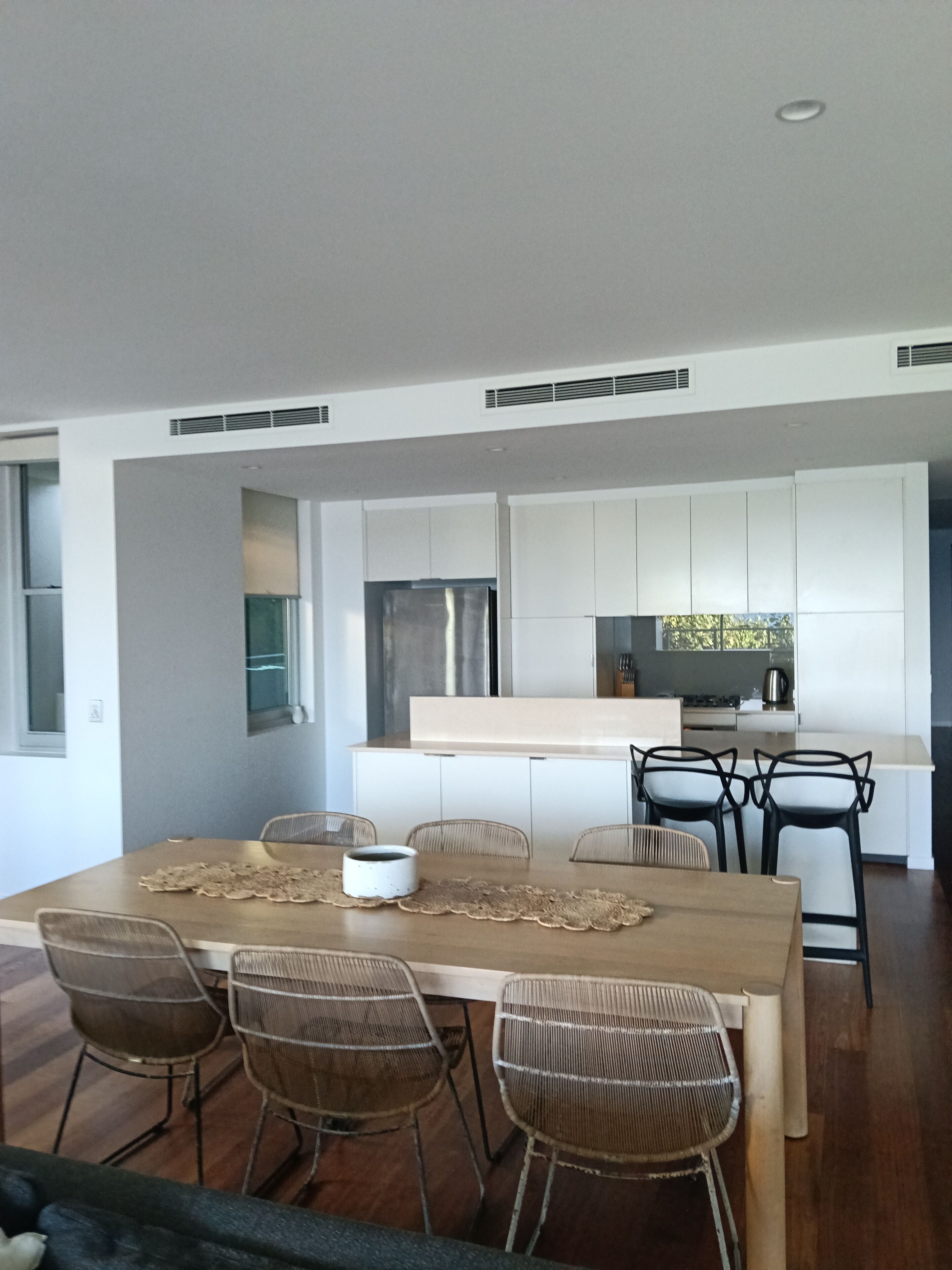 Une cuisine moderne avec des meubles blancs, un îlot central et un coin repas comprenant une table en bois et des chaises en osier.