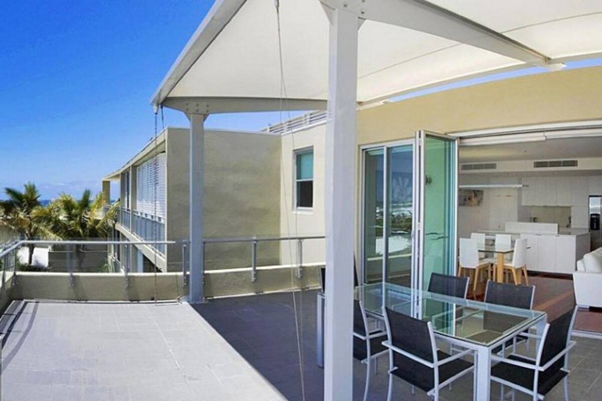 A modern outdoor patio with a glass table and chairs, a covered seating area, and a view of the ocean.