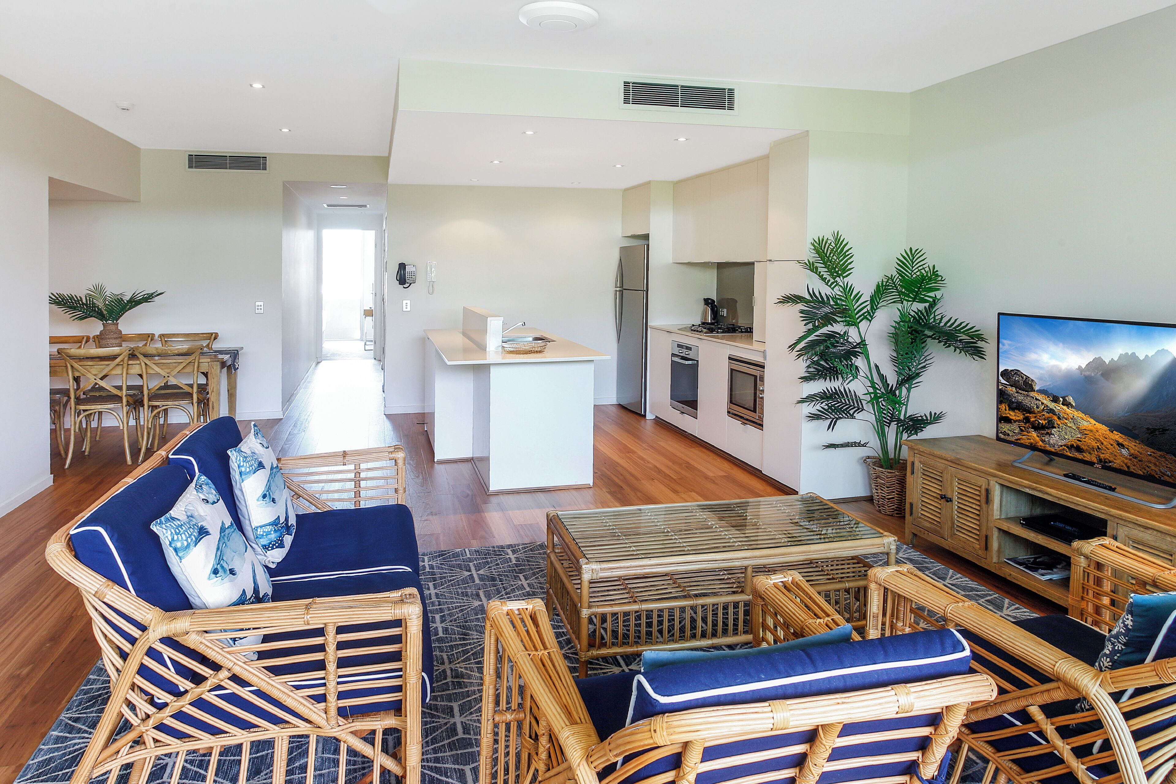 A modern living room with wicker furniture, a dining area with wooden chairs, a kitchen with a refrigerator and stove, and a TV on a wooden cabinet.