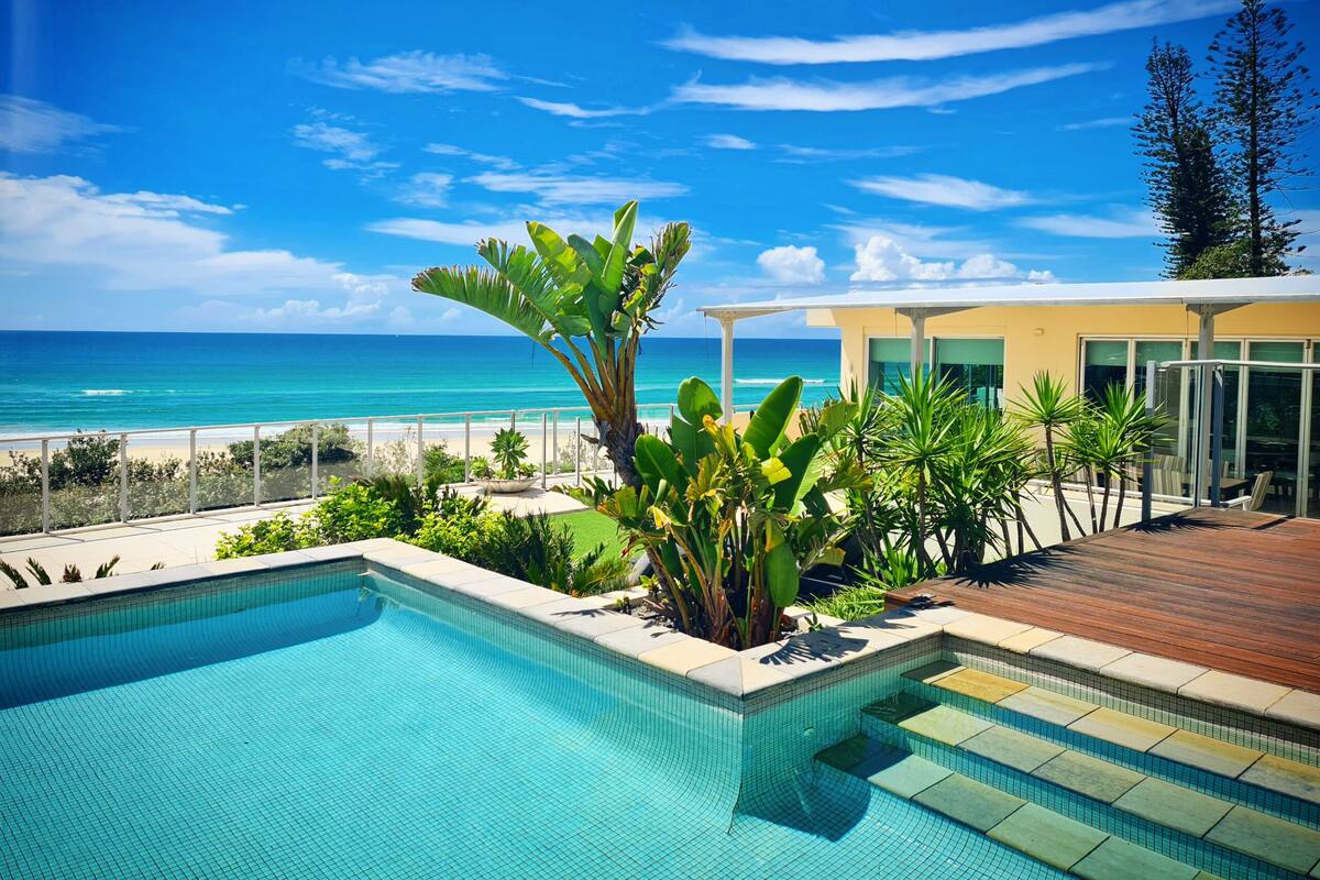 A pool with a view of the ocean, surrounded by tropical plants and a wooden deck.