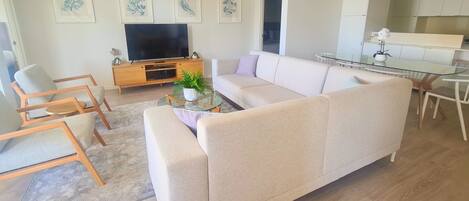 A modern living room with a beige sofa, a glass coffee table, and a flat-screen TV. There are framed artworks on the wall and a small plant on the coffee table.