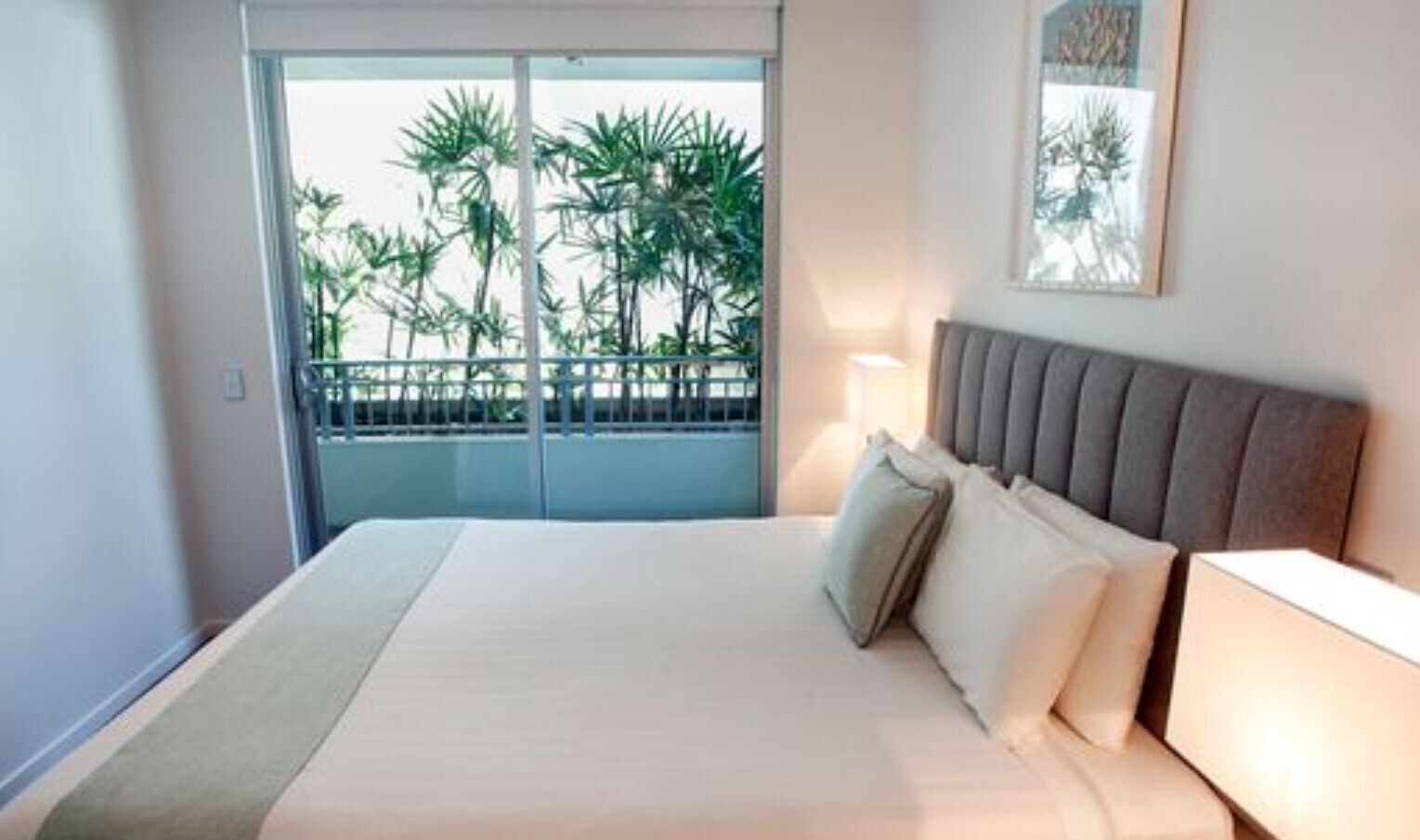 A hotel room with a bed, a nightstand, a lamp, a balcony with a view of palm trees, and a framed picture on the wall.