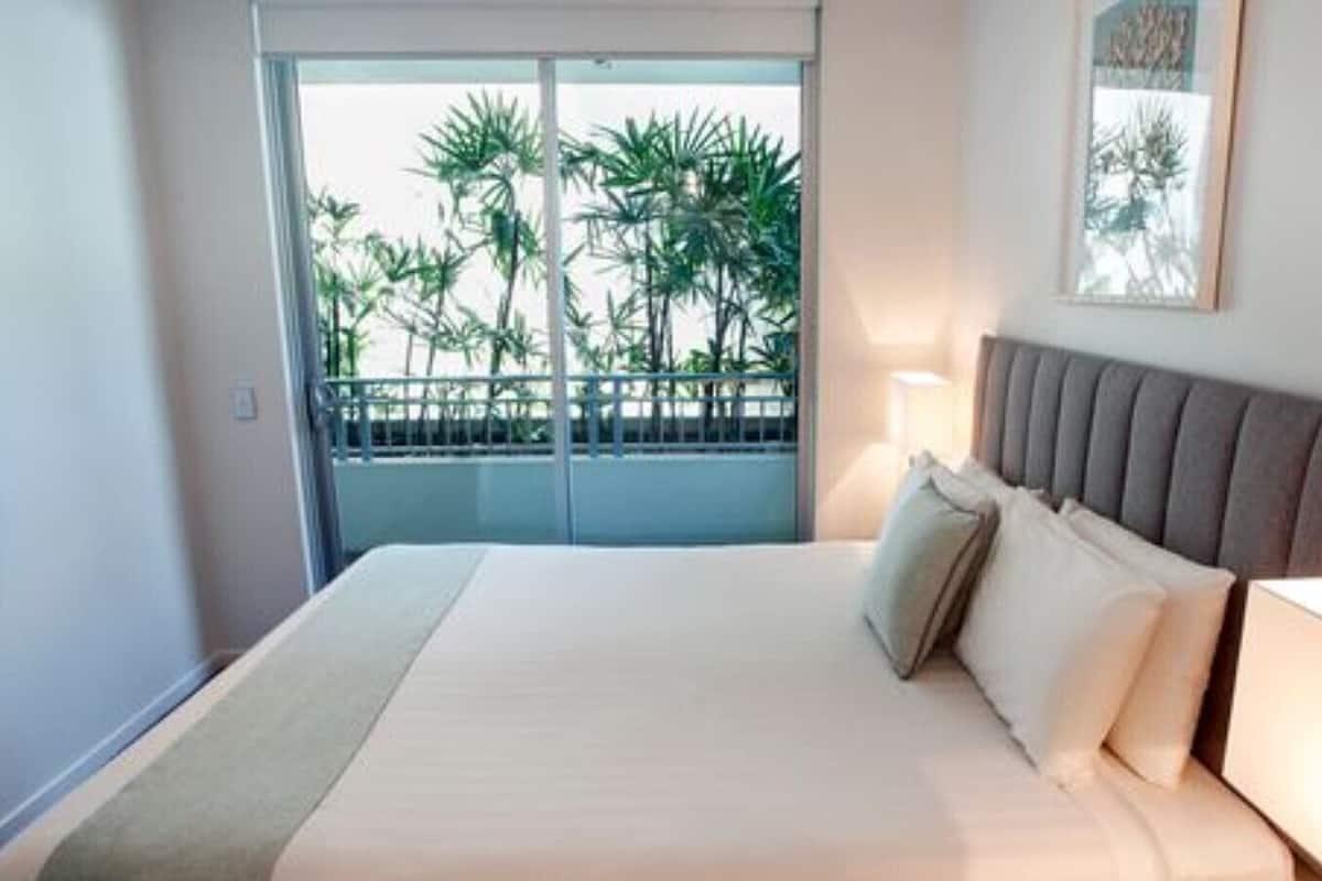 A hotel room with a bed, a nightstand, a lamp, a balcony with a view of palm trees, and a framed picture on the wall.