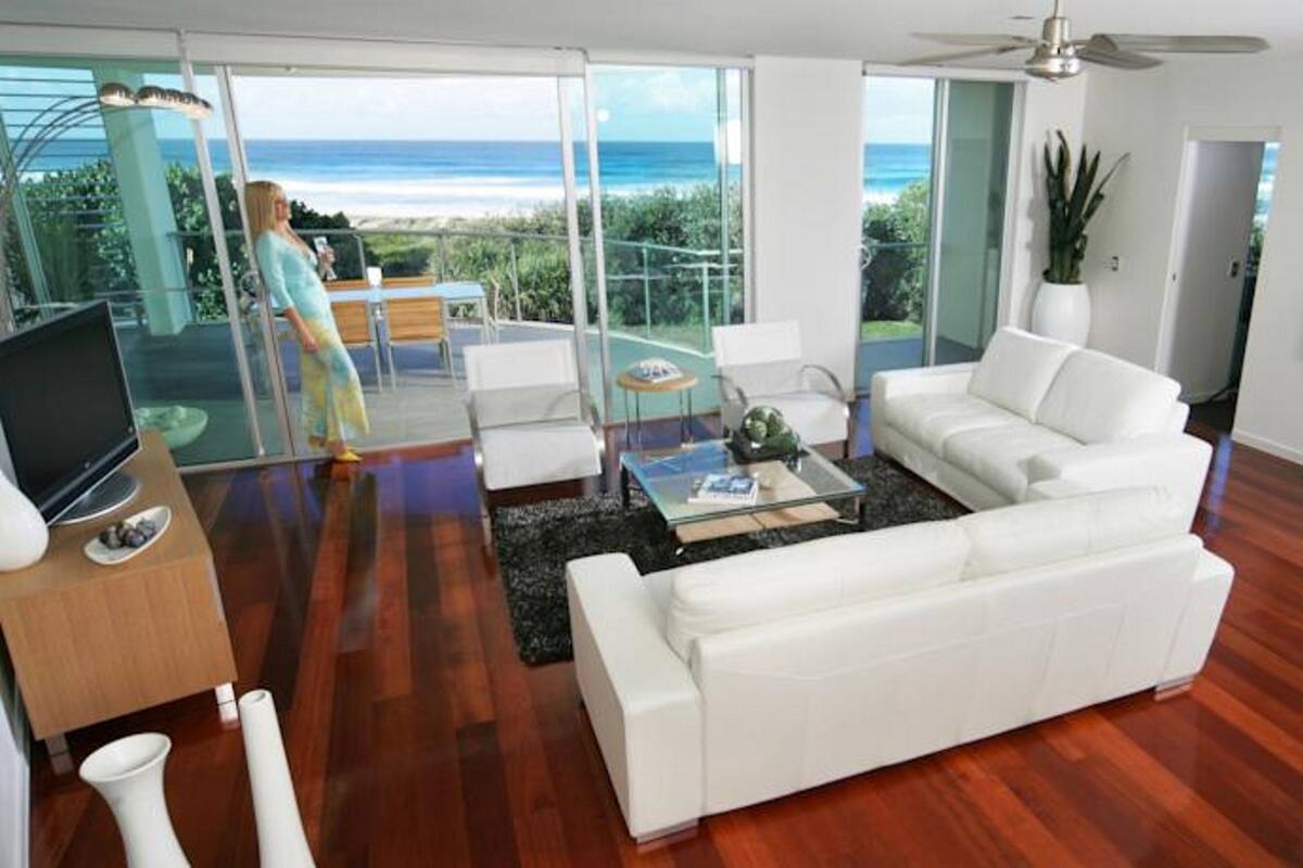 A modern living room with a white sofa, a glass coffee table, and a TV on a wooden stand. There is a balcony with a view of the ocean.