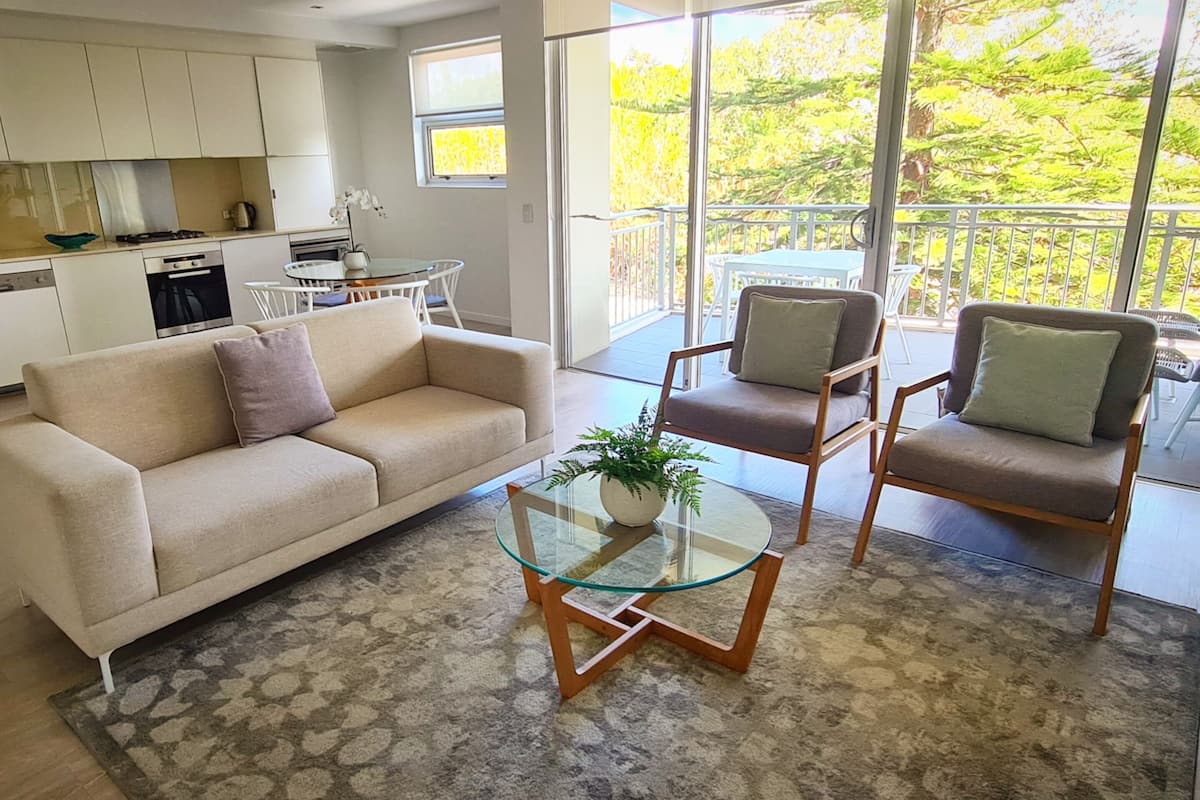 A modern living room with a sofa, two armchairs, a glass coffee table, and a view of a balcony with outdoor furniture.