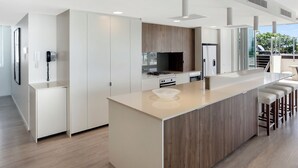 A modern kitchen with a central island, bar stools, a built-in oven, and a large window offering a view of the outdoors. - The Beach Cabarita (Cabarita Beach)