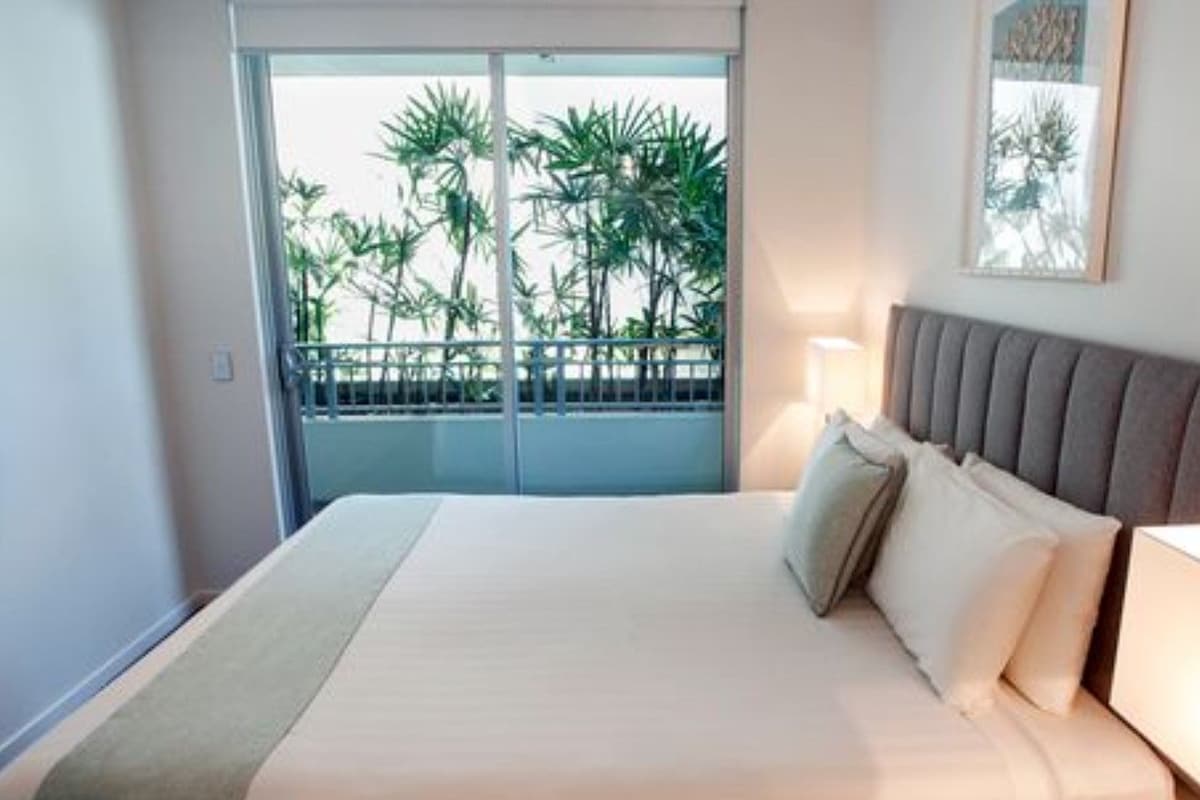 A hotel room with a bed, a nightstand, a lamp, a balcony with a view of palm trees, and a framed picture on the wall.