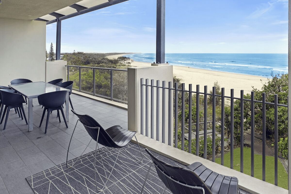 Ein Balkon mit Tisch und Stühlen, der Blick auf einen Strand und das Meer freigibt.