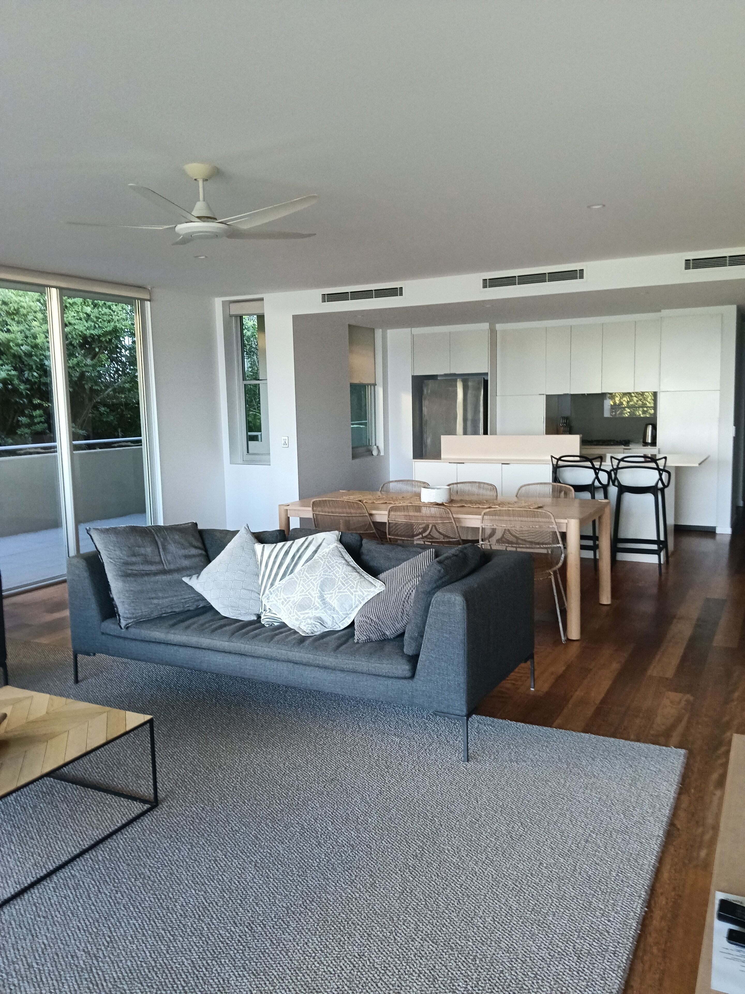 A modern living room with a grey sofa, a wooden dining table, and a kitchen in the background.