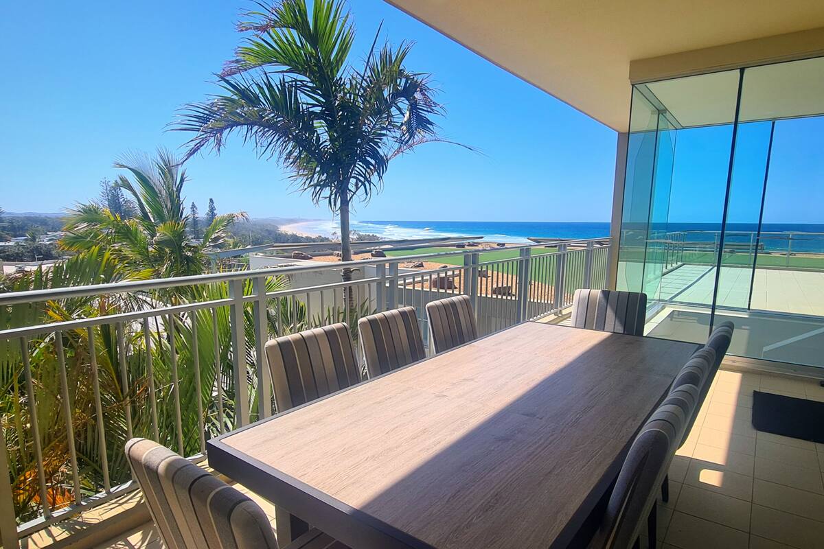 A balcony with a wooden table and chairs, overlooking a beach and ocean.
