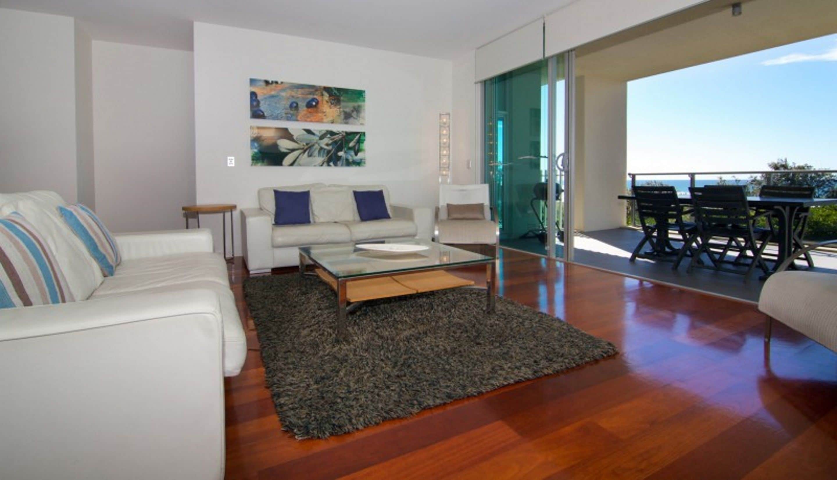 Un salon moderne avec un canapé blanc, une table basse en verre et une grande fenêtre donnant sur l’extérieur.