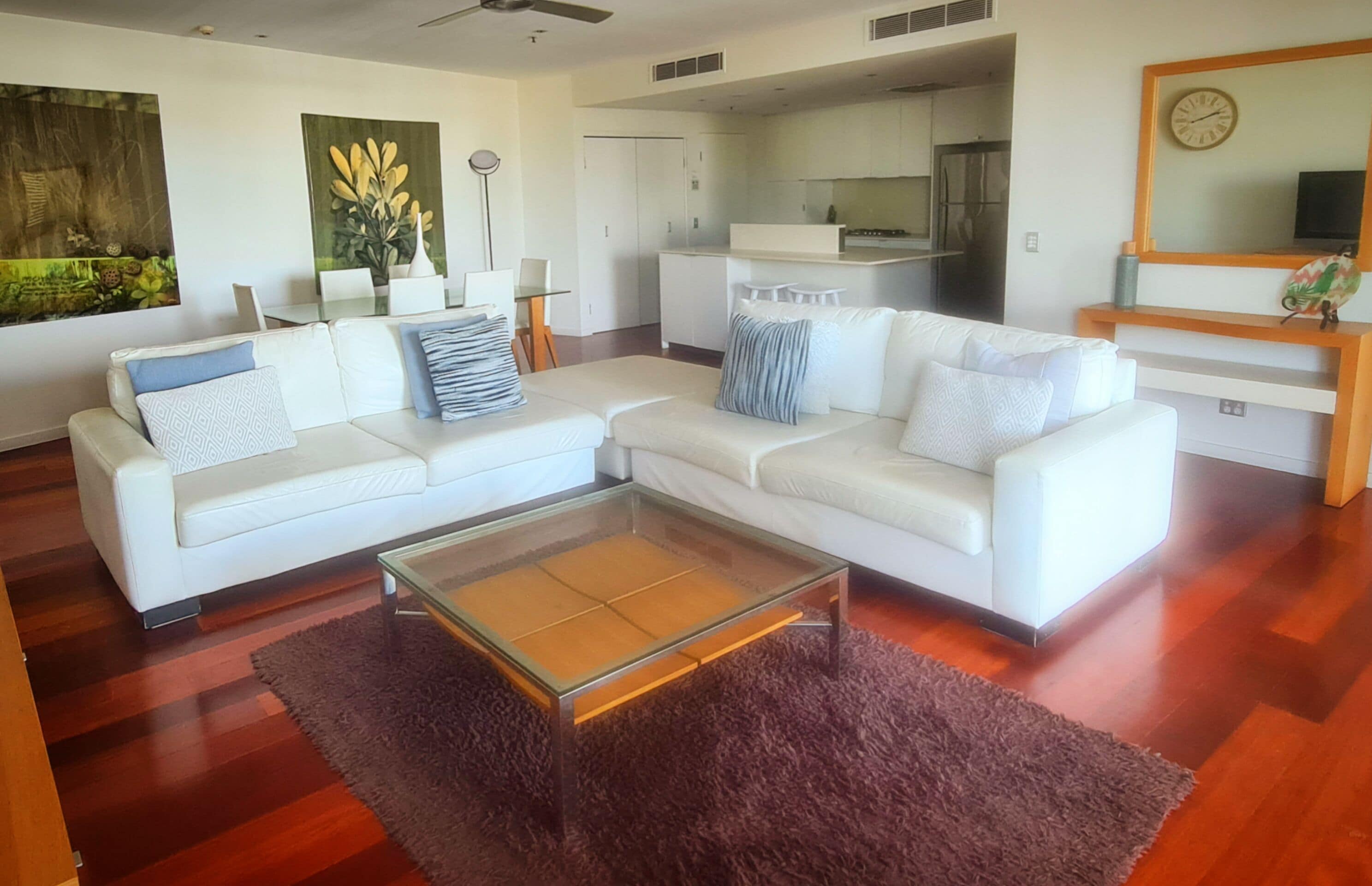 A modern living room with a white sectional sofa, a glass coffee table, and a kitchen in the background.