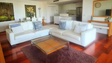 A modern living room with a white sectional sofa, a glass coffee table, and a kitchen in the background.